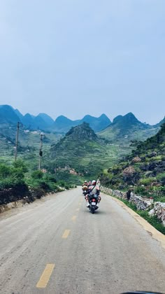 a motorcycle is driving down the road in front of some mountains and grass on either side