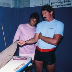 two men standing in front of a surfboard with the board on it's end