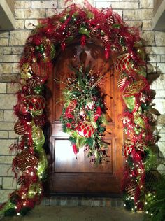 a door decorated with red and green christmas wreaths next to a brick wall covered in greenery