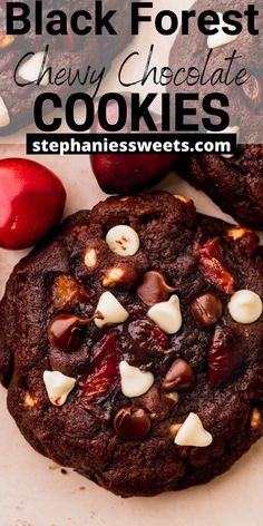 a chocolate cookie with white chocolate chips and cherries on top, next to an apple