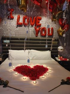 a bed topped with lots of red roses and candles next to balloons that spell i love you