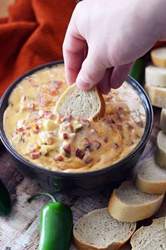 a hand dipping a piece of bread into a bowl filled with cheese and bacon dip