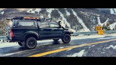 a black truck driving down the road with snow on the mountains in the back ground