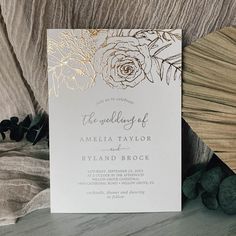 an elegant wedding card with gold foil flowers on the front and back, sitting next to some rocks