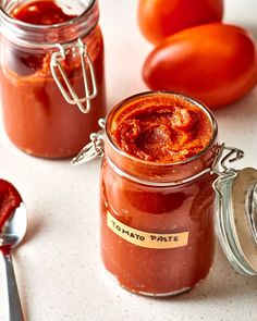 two jars filled with tomato paste next to tomatoes and spoons on a white surface