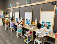 two children are sitting at desks with easels and paintings on the wall behind them