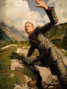 a woman with her hand up in the air while standing on rocks and grass near mountains