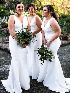 three bridesmaids in white dresses posing for the camera with greenery around them