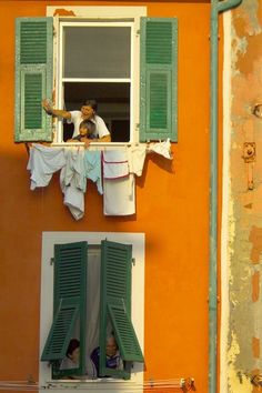 two people are looking out the window of an orange building with green shutters and drying clothes