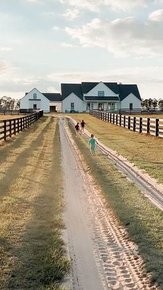 two people walking down a dirt road in front of a white house