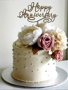 a white cake with pink flowers on top and happy anniversary written on the side in cursive writing