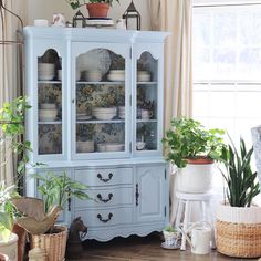 a blue china cabinet with plates and plants on top in a living room area next to a window