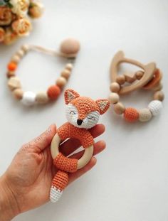 a hand holding an orange knitted fox toy in front of some beads and flowers