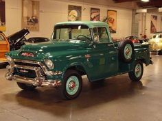 an old green pickup truck parked in a garage with other classic cars on display behind it