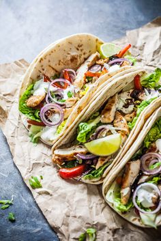 three chicken tacos with lettuce, tomatoes and onions on parchment paper next to lime wedges