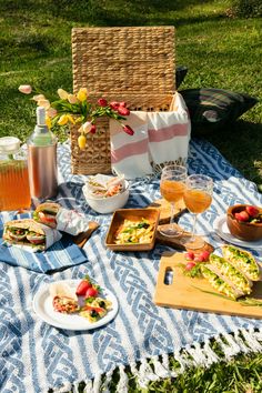 an outdoor picnic with food and drinks on the blanket in the grass, next to a wicker basket full of flowers