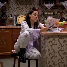 a woman sitting on a stool reading a paper in front of a counter with fruit