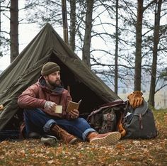 a man sitting in front of a tent reading a book next to a bag and backpack