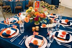 the table is set with blue linens, orange and white place settings, silverware, and flowers