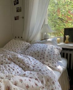 an unmade bed in front of a window with white curtains and flowers on it