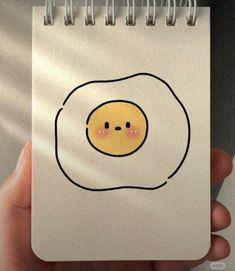 a hand holding a spiral notebook with an egg drawn on it