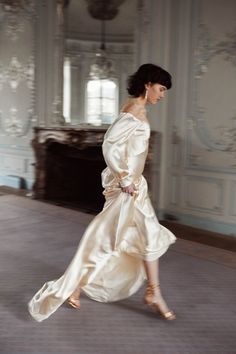 a woman in a white dress walking across a room with an ornate wall behind her