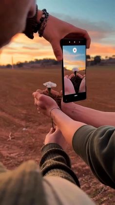 two people holding up their cell phones in the middle of an open field at sunset