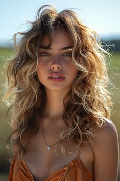 Close-up of a woman with sunlit tousled hair, wearing an orange top and a heart-shaped pendant, gazing at the camera. Fairy Kissed Hair, Sun Kissed Hair Brunette, Balayage Styles