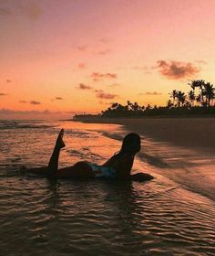 a woman laying on her stomach in the ocean at sunset with palm trees behind her