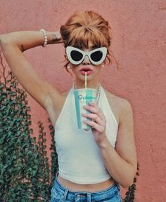 a woman wearing sunglasses and holding a drink in front of her face while standing next to a pink wall