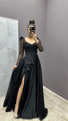 a woman taking a selfie wearing a black dress with sheer sleeves and thigh high slit