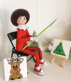 an elf sitting in a chair holding a paintbrush next to a small christmas tree