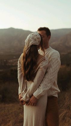 a man and woman standing in the middle of a field with mountains in the background