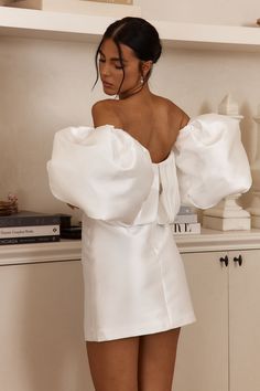 a woman standing in front of a book shelf wearing a white off the shoulder dress