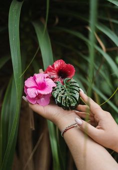 a person holding flowers in their hand with palm leaves around them and one pink flower on the other side