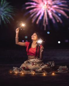 a woman sitting on the ground holding sparklers in her hand and looking up at fireworks