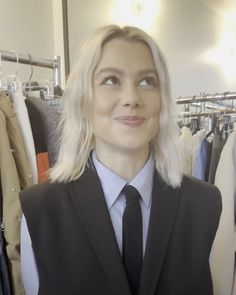 a woman with blonde hair wearing a suit and tie in front of clothes on hangers