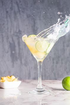 a martini glass filled with lemonade and limes on a table next to a bowl of chips