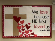 Brown paper cross, some sparkly red and some red construction paper hearts coming from the center of the cross and spilling to the bottom of the bulletin board. Text is “We love because he first loved us. 1 John 4:19”.