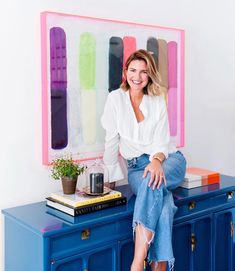 a woman sitting on top of a blue dresser in front of a colorful art piece