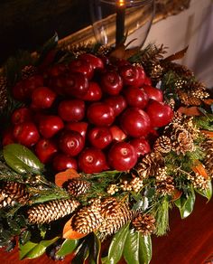 a bowl filled with apples and pine cones on top of a wooden table next to a candle