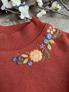 a red sweater with flowers and leaves embroidered on the front, sitting next to white flowers