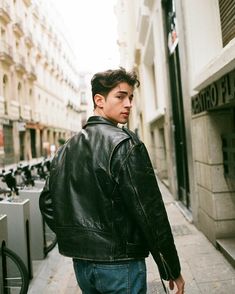 a young man walking down the street with his hand in his pocket and wearing a leather jacket