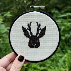 a hand holding up a cross - stitched deer ornament with red eyes