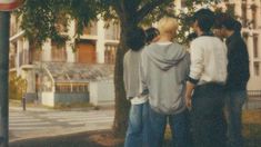 four people standing in front of a tree on the side of the road with buildings behind them