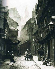 an old black and white photo of people sitting on the sidewalk in front of buildings