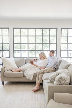 a man and woman are sitting on a couch in front of large windows, while holding each other