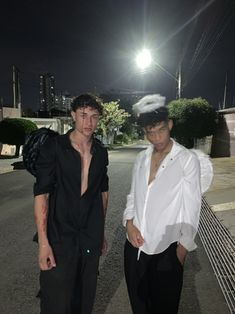 two young men standing next to each other on a street at night with lights shining in the background