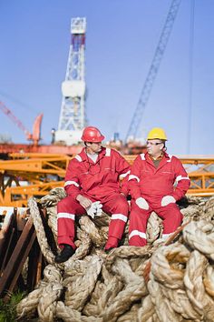 two men in red work suits sitting on top of rope next to a large crane