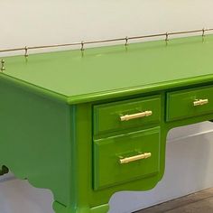 a green desk with gold handles on the top and two drawers at the bottom, in front of a white wall